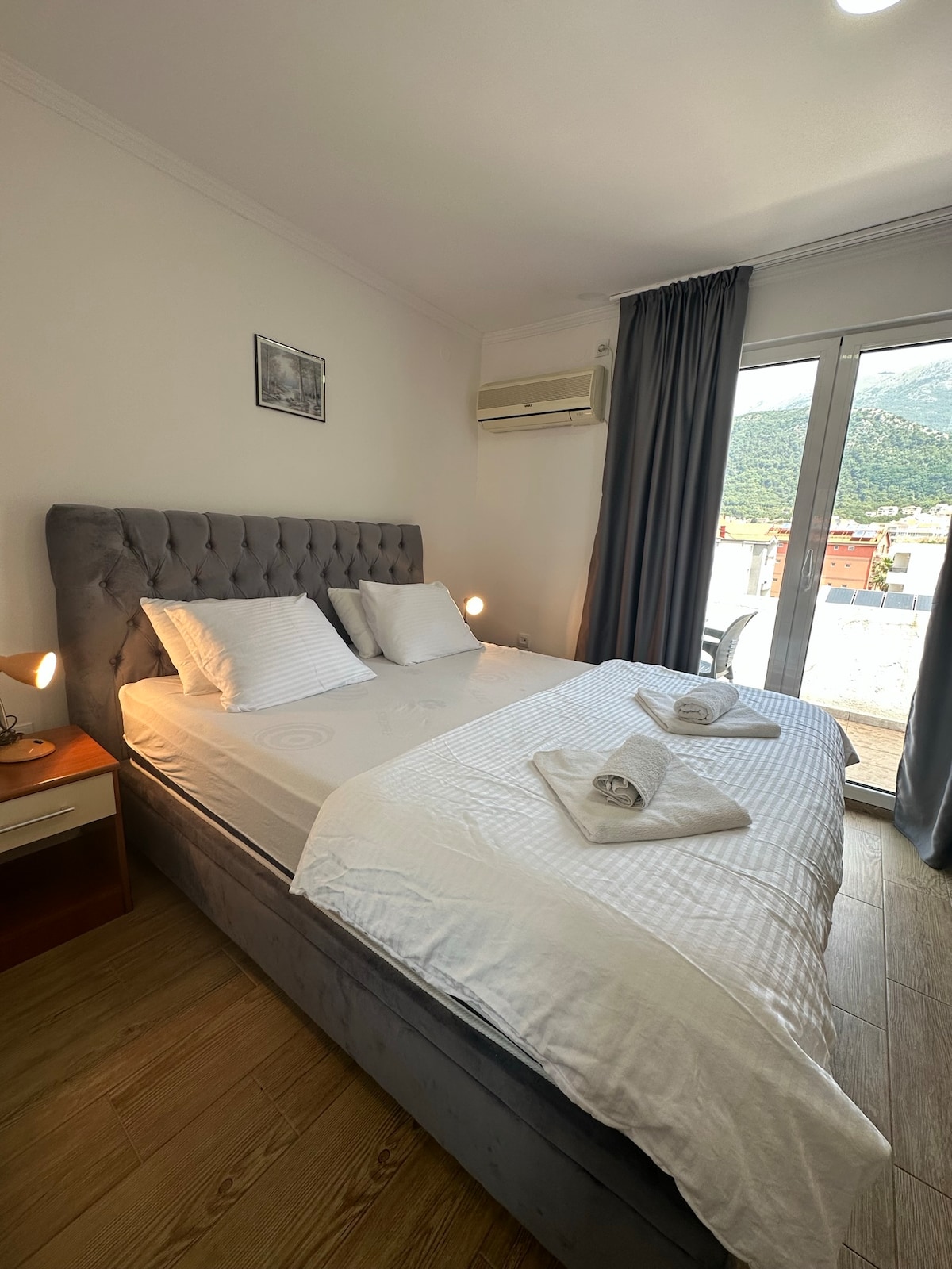 A well-furnished bedroom features a spacious bed with neatly arranged linens and two towels. Natural light enters through large glass doors that open to a view of the surrounding hills, while soft grey curtains frame the windows.