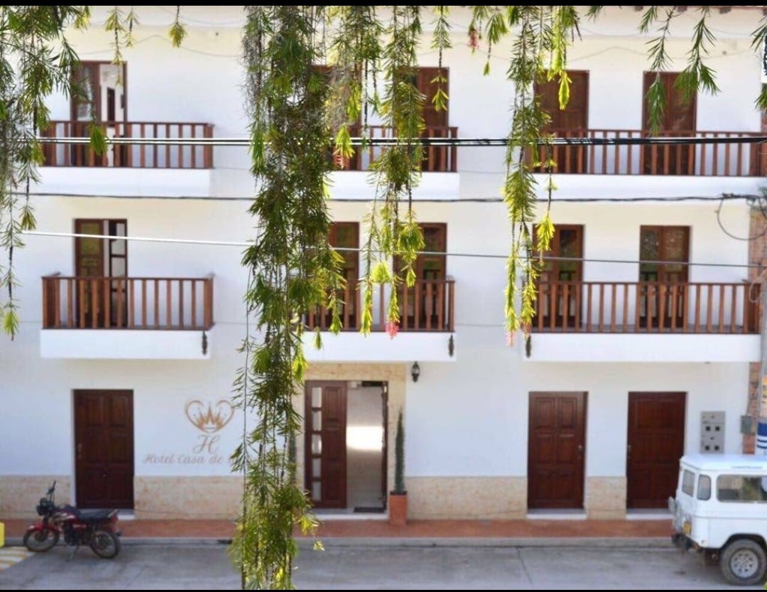 A charming hotel exterior is presented with a row of wooden doors leading to the entrance. Balconies with wooden railings are visible on the upper floors, while a motorcycle and a vintage vehicle are parked nearby. Lush greenery hangs from above, adding a natural element.