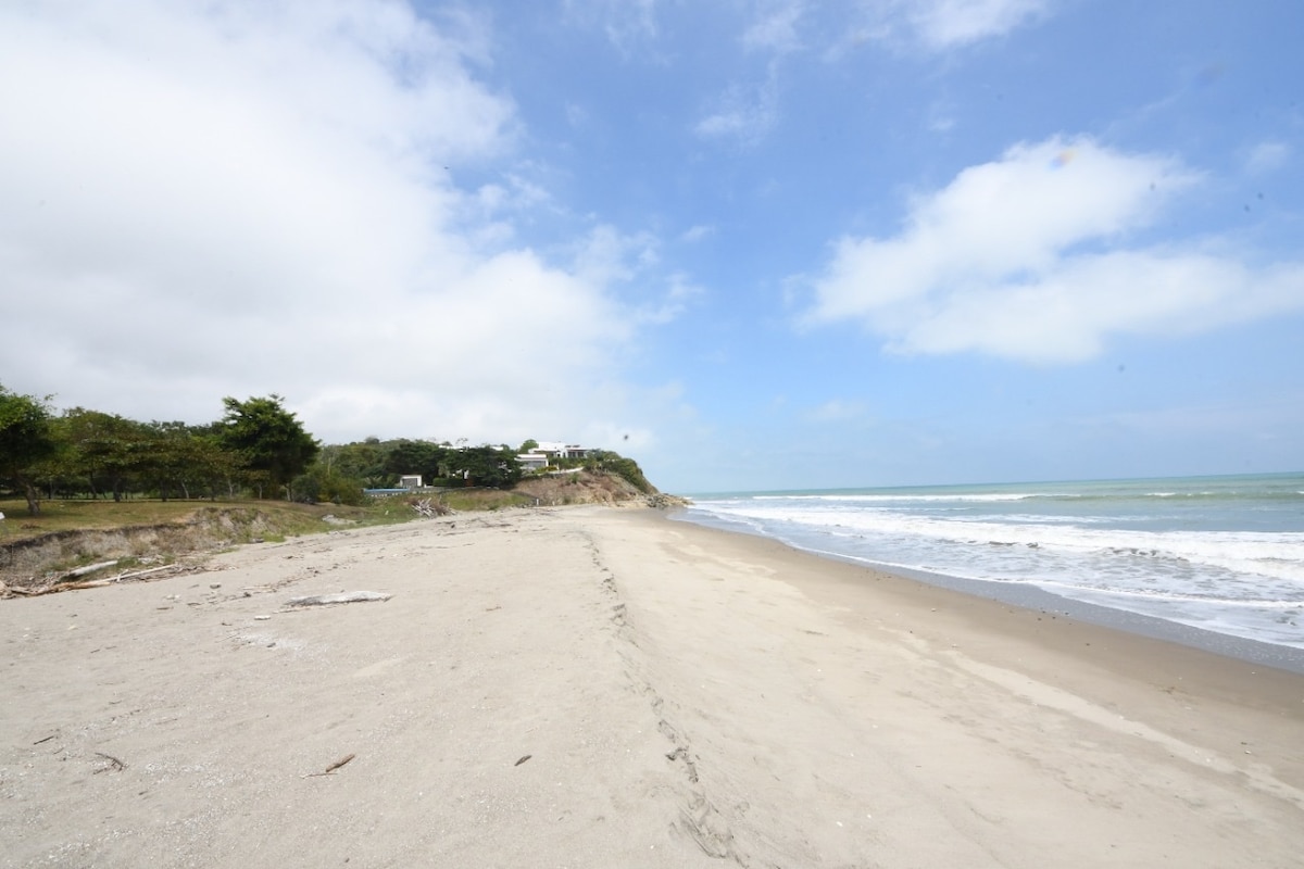 A sandy beach is seen stretching along the shoreline, with gentle waves lapping at the edge. The sky is partly cloudy, creating a serene atmosphere. A distant cliff is visible in the background, providing a natural backdrop to the tranquil scene.