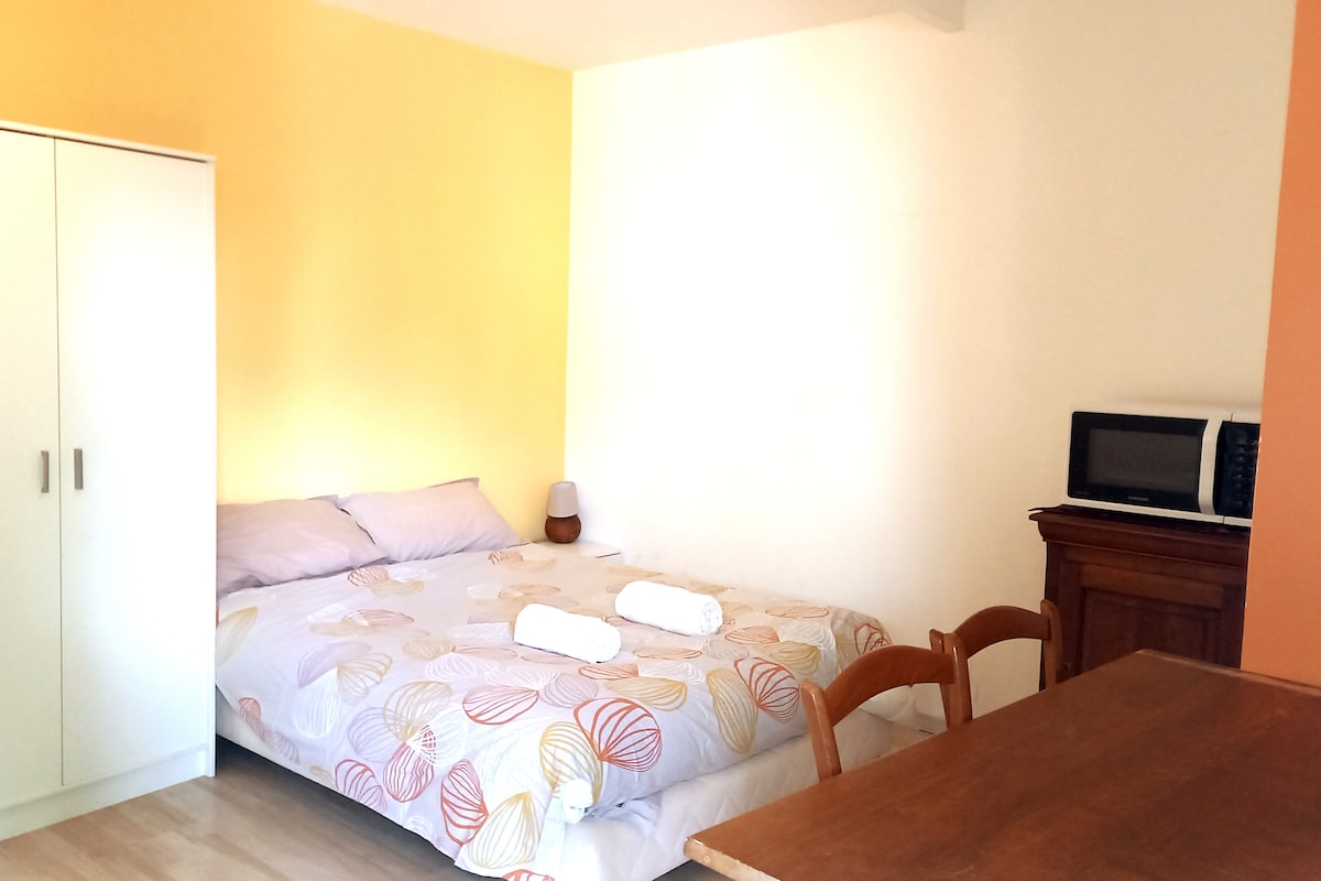 An open studio features a spacious bed with a patterned coverlet, flanked by two towels. A wooden dining table with chairs occupies one corner. A wardrobe stands against the wall, while a small microwave is positioned nearby. The walls are painted in soft yellow and orange hues.