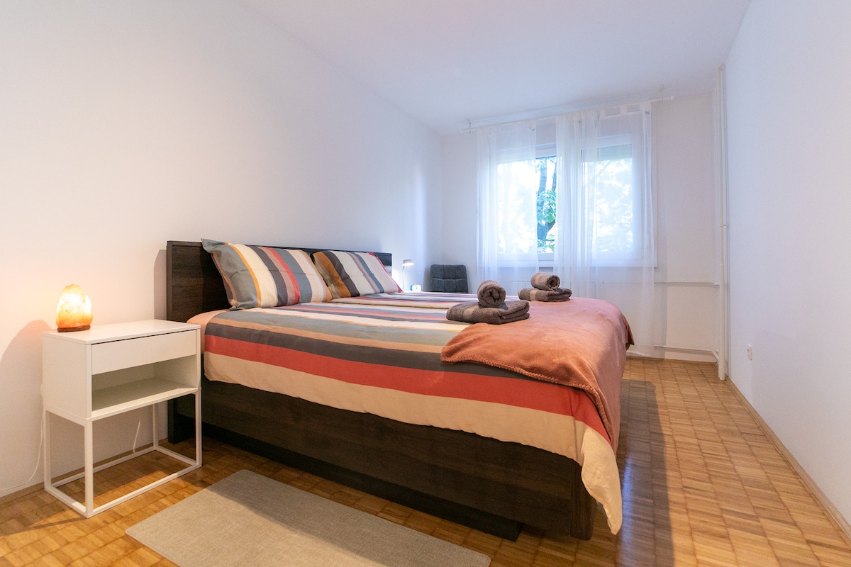 A spacious bedroom features a large bed with striped bedding and soft towels arranged neatly. Natural light filters in through a window, complementing the warm tones of the room. A small nightstand with a lamp sits beside the bed, while a comfortable chair is positioned in the corner.