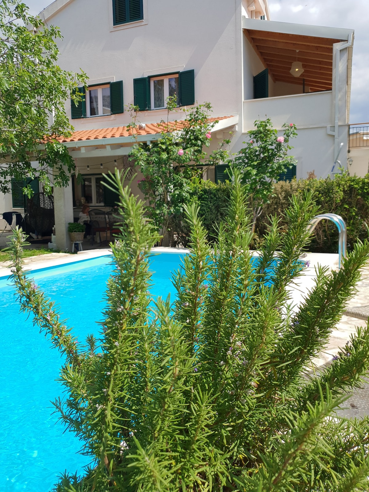 A refreshing swimming pool is framed by vibrant greenery, with a well-maintained garden surrounding the area. The spacious villa, featuring green shutters and a terracotta roof, is visible in the background, complemented by blooming plants and shaded outdoor seating.