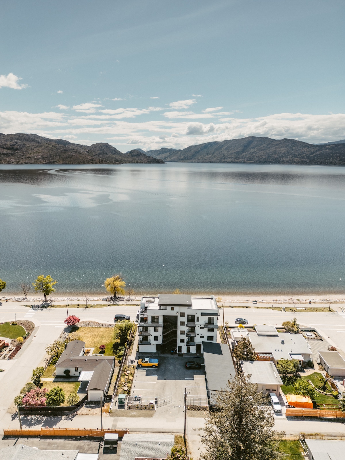 A stunning aerial view captures the shoreline of Okanagan Lake, with mountains in the background. The apartment building is positioned adjacent to a sandy beach, framed by lush greenery and colorful trees. Clear blue skies reflect on the water's surface, enhancing the serene atmosphere.
