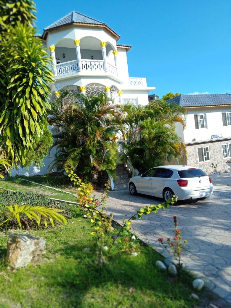 A three-story home is surrounded by lush greenery and tropical plants. A vehicle is parked on a paved driveway leading to the entrance. The architecture features a balcony with decorative railings and a bright exterior with yellow accents against a clear blue sky.