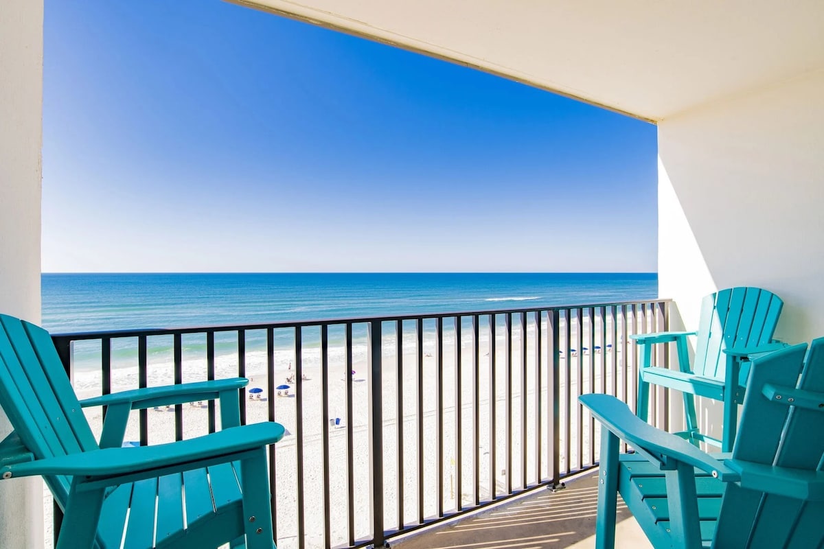 A balcony with bright turquoise chairs provides a panoramic view of the beach and ocean. The horizon stretches under a clear blue sky, while the inviting sandy shore is visible below, enhancing the connection between indoor and outdoor spaces.