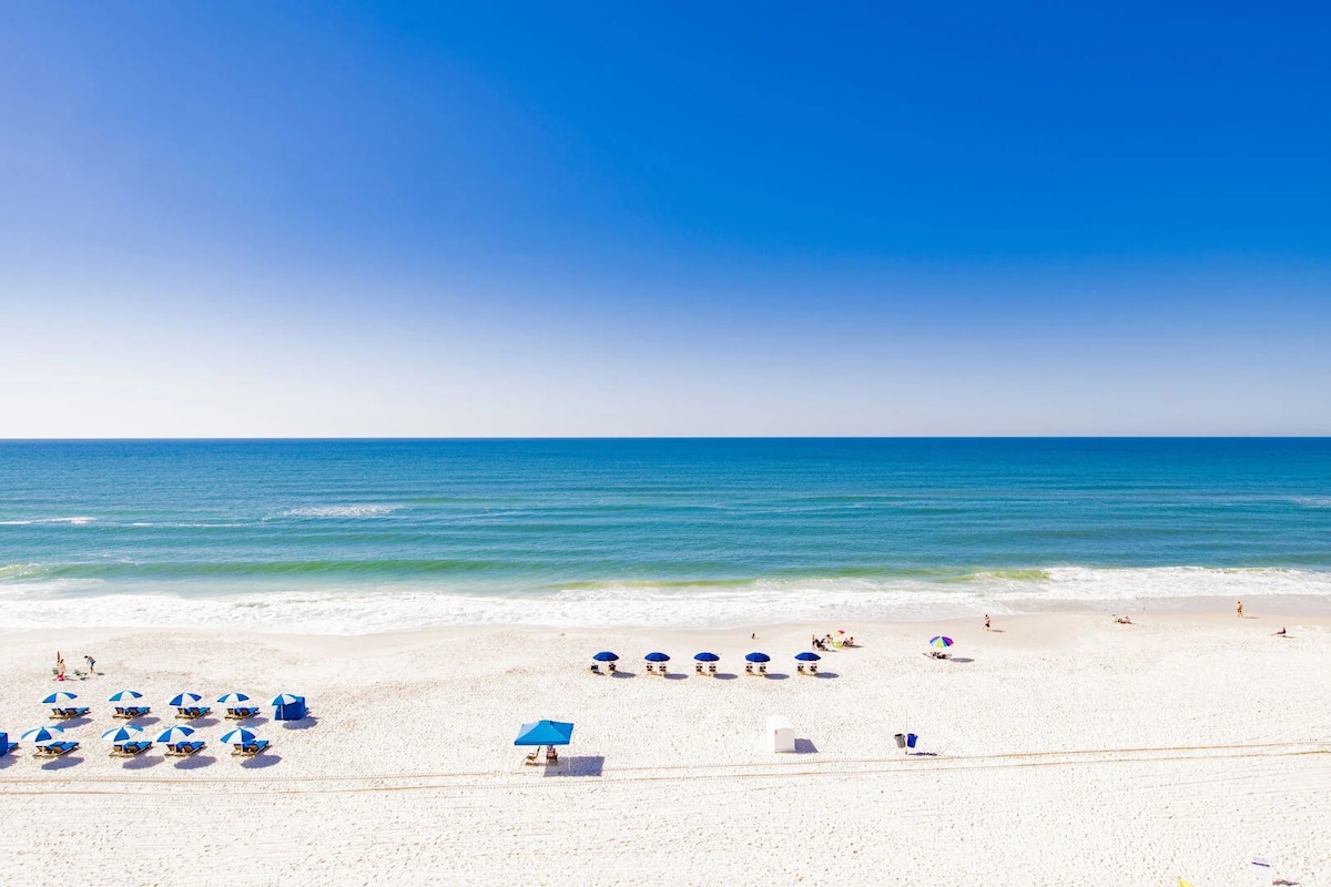 A panoramic view of the beach showcases soft white sand meeting clear blue waters, with several blue umbrellas and lounge chairs arranged on the shore. The horizon blends seamlessly with the sky, creating a serene coastal scene.