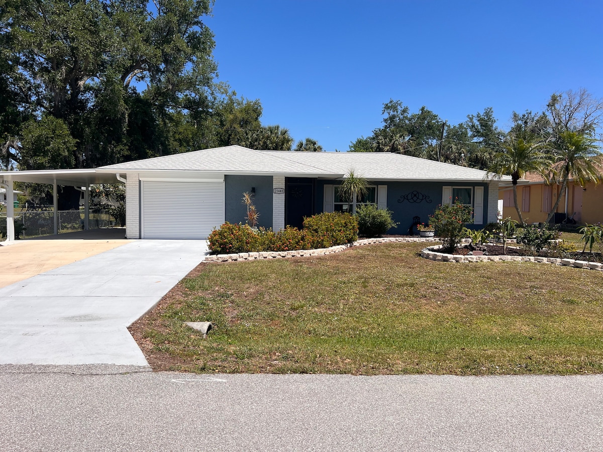 The exterior of the home features a single-story structure with a light-colored roof and a spacious driveway. Decorative landscaping includes a variety of shrubs and palm trees, creating a welcoming entrance. The clear blue sky enhances the bright atmosphere of the surrounding area.
