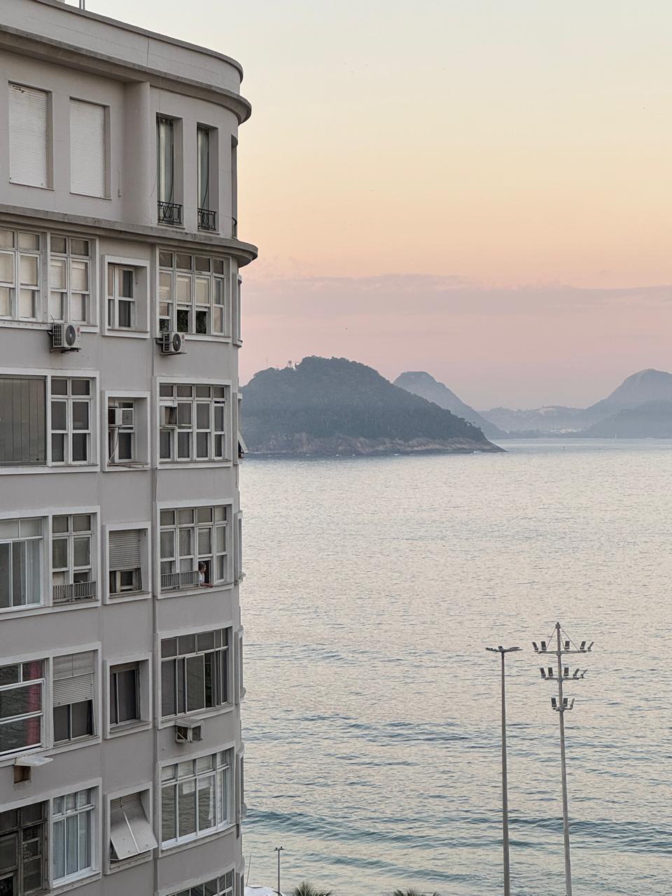 The image captures the corner of a light-colored apartment building, with large windows visible. Beyond the building, the serene ocean is visible, reflecting soft pastel hues of the sky at dusk. Gentle hills rise in the distance, adding to the tranquil coastal scene.