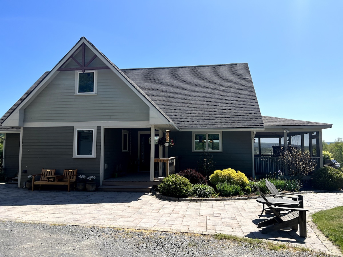 The exterior of a welcoming house features a combination of gray and white siding beneath a sloped roof. A spacious porch is adorned with a wooden railing, while seating areas and colorful landscaping enhance the inviting entrance. A clear blue sky completes the serene setting.
