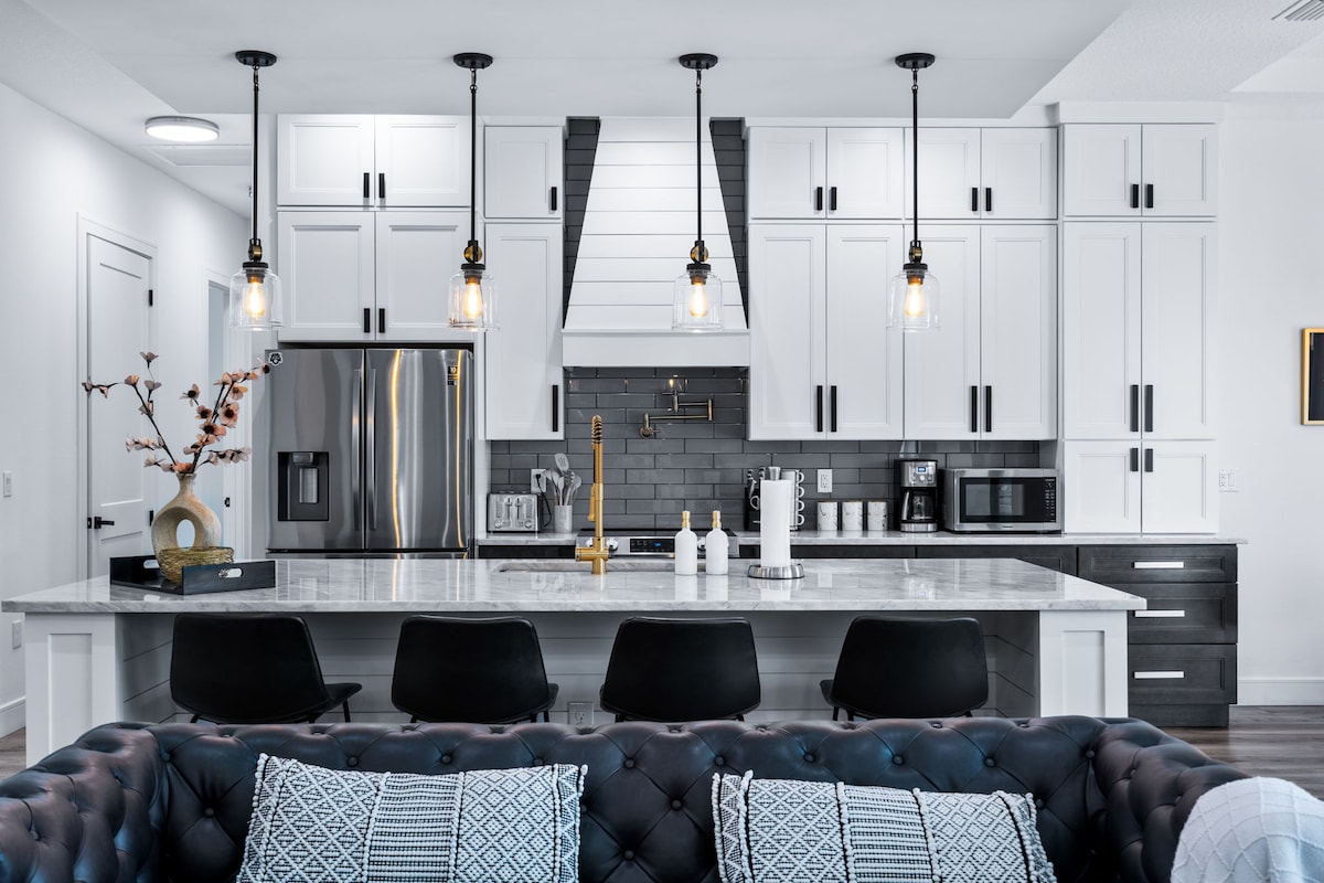 The kitchen area features a centralized island with bar seating, complemented by modern pendant lighting. A sleek refrigerator and stainless steel appliances are visible, alongside dark cabinetry and a stylish backsplash. A cozy sitting area with a tufted couch offers a warm gathering space.