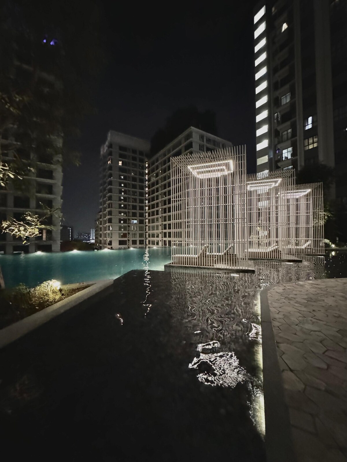 A modern pool area is observed, highlighted by illuminated architectural features reflecting in calm waters. Surrounding buildings showcase a sleek design, with soft lighting accentuating the water's surface at night.