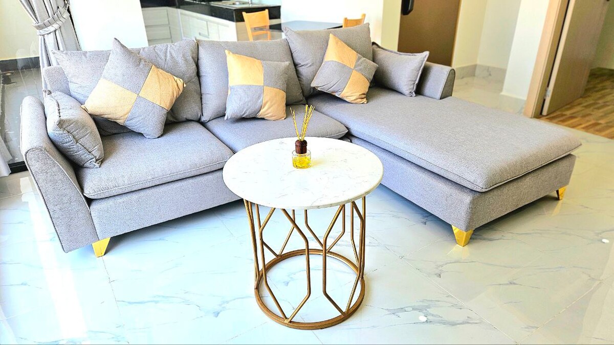 A modern sectional sofa in light grey is accented with golden square cushions. A marble-top coffee table with a gold geometric base is positioned in front of the sofa, creating a comfortable and stylish living area, illuminated by natural light from nearby windows.