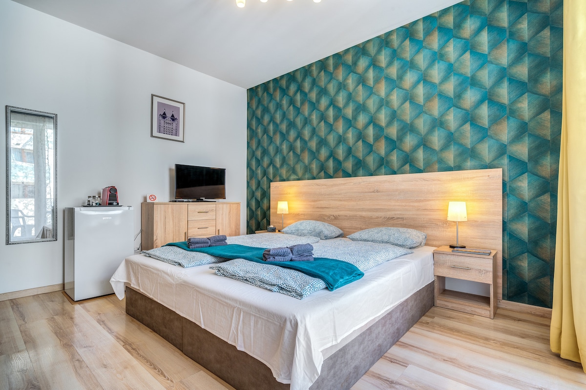 A spacious bedroom features a geometric patterned accent wall in calming turquoise. A large bed with soft linens and neatly arranged towels is paired with a wooden nightstand and lamp. A modern dresser and a small refrigerator are visible, along with a flat-screen television.