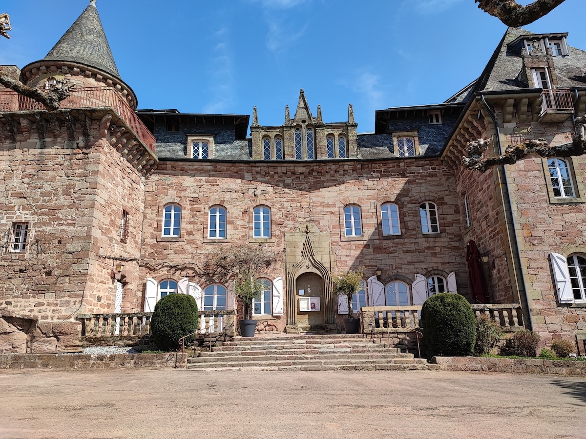 The historic façade of a castle features intricately designed stonework with a mix of towers and arched windows. Symmetrical staircases lead to a central entrance, framed by manicured shrubs and stone balustrades, all set against a clear blue sky.