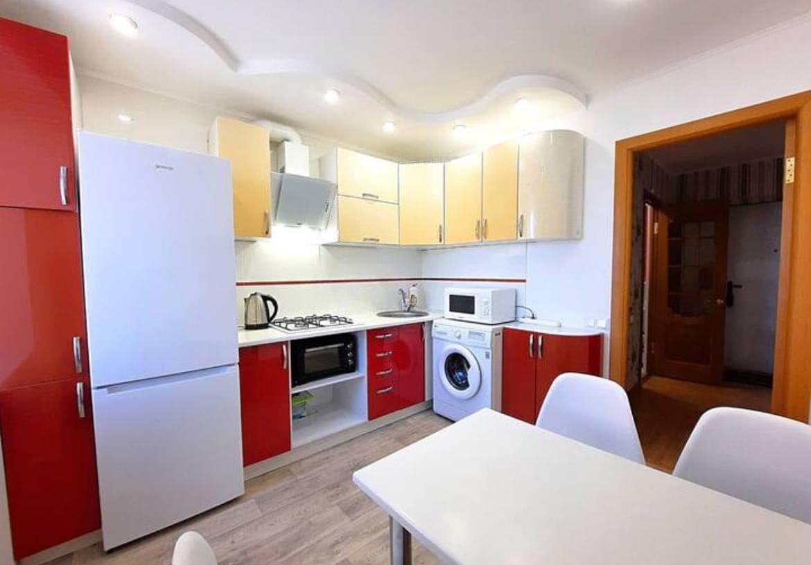 A modern kitchen is equipped with red and cream cabinetry. Stainless steel appliances, including a refrigerator, stove, and microwave, are present. A white dining table with gray chairs is positioned in the center, and a washing machine is located near the counter.