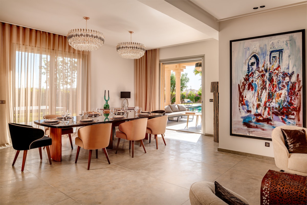 An elegant dining area features a long, dark wooden table surrounded by plush, upholstered chairs in warm tones. Two crystal chandeliers hang from the ceiling. Floor-to-ceiling windows invite natural light and offer views of the outdoor terrace and pool area.