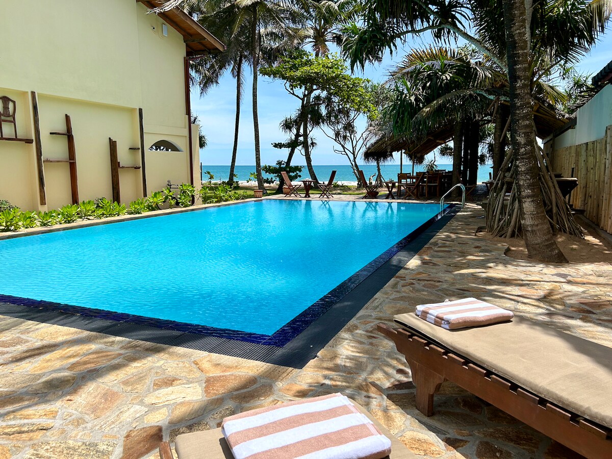 A large private pool is framed by lush palm trees, providing direct views of the ocean. Comfortable lounge chairs with striped towels are positioned around the pool, and natural stone tiles create a seamless connection to the surrounding tropical landscape.