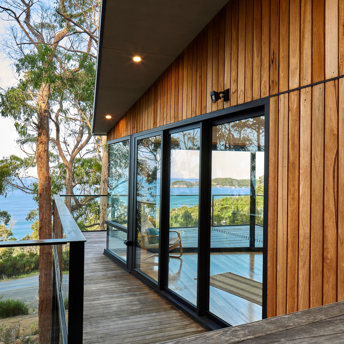 An outdoor deck extends from the modern home, featuring glass sliding doors that offer unobstructed views of Pirates Bay. The surrounding trees enhance the natural setting, while a small seating area is visible on the deck, inviting relaxation in a serene environment.