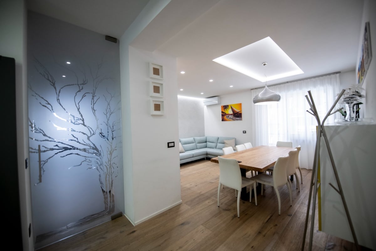An open-plan living area is visible, featuring a light wooden floor. A spacious, modern sofa is situated against the wall, complemented by a large dining table surrounded by white chairs. Natural light flows in through sheer curtains, enhancing the room's bright ambiance.