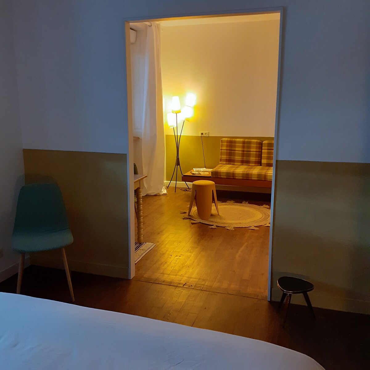 A view from the bedroom opens into a cozy sitting area with a checked sofa. Soft lighting is provided by a trio of standing lamps, enhancing the warm ambiance. A small round table is positioned beside a textured rug, creating an inviting space for relaxation.