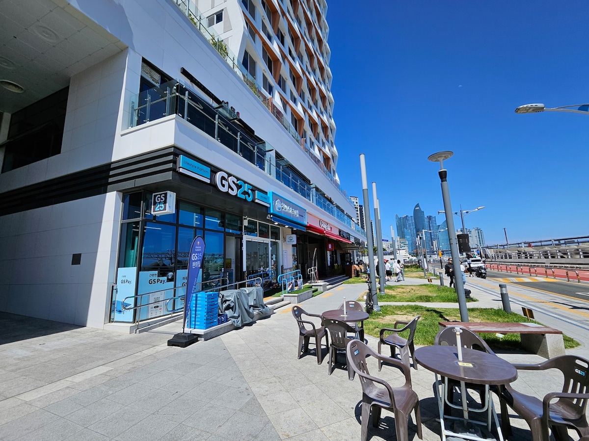 A vibrant streetscape is depicted, featuring a modern building with a mix of shops and eateries on the lower level. Outdoor seating is available with several tables and chairs arranged on the tiled area, while a clear blue sky enhances the scene.