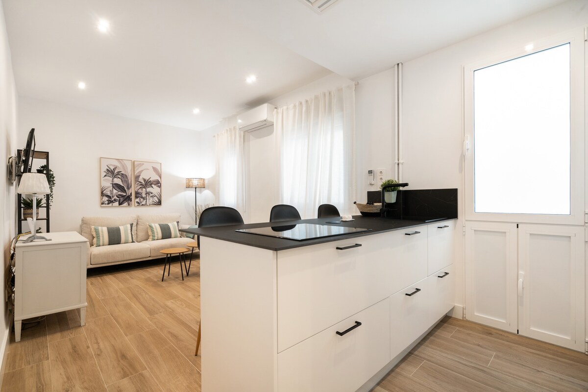 A bright and airy kitchen opens to a cozy living area, featuring a modern island with seating. Large windows allow natural light to fill the space, complemented by light wood flooring. The inviting seating area includes a comfortable sofa and a small coffee table.