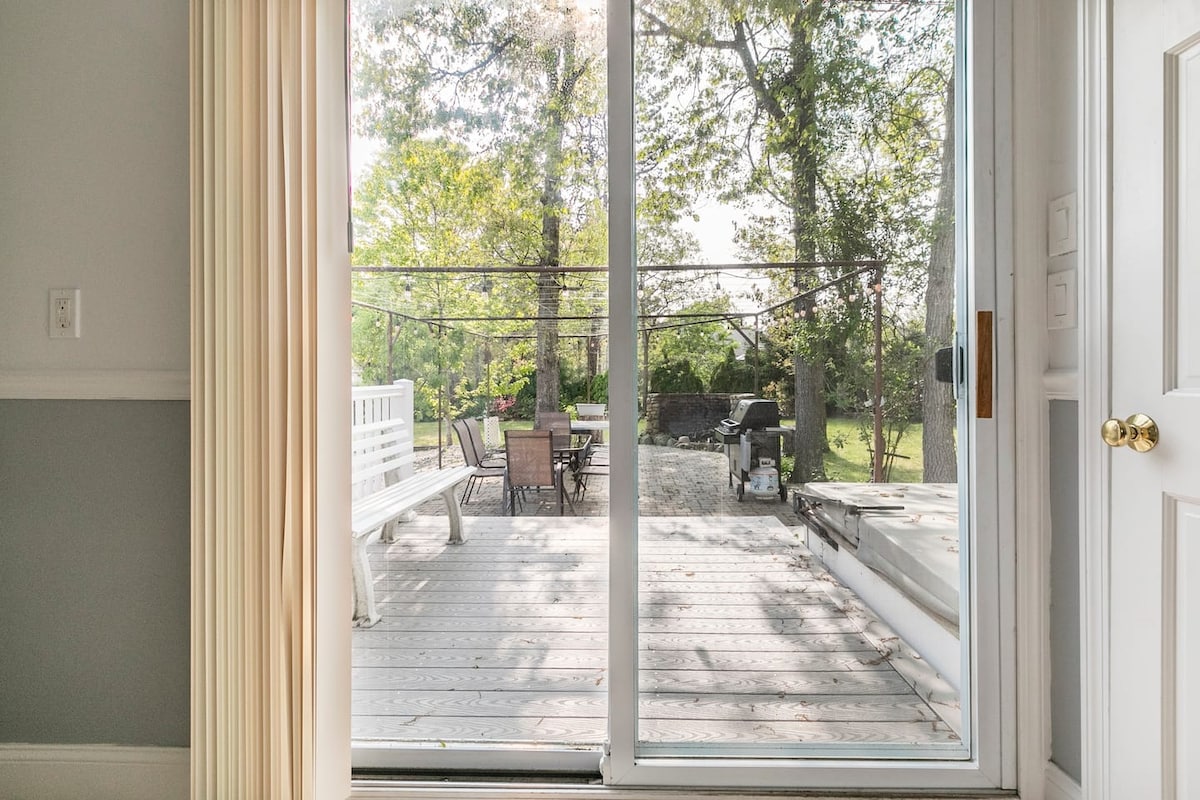 A large sliding glass door opens to an outdoor deck surrounded by green trees. The deck features a picnic table and additional seating, providing space for relaxation and dining. A glimpse of a spacious yard is visible beyond the deck.
