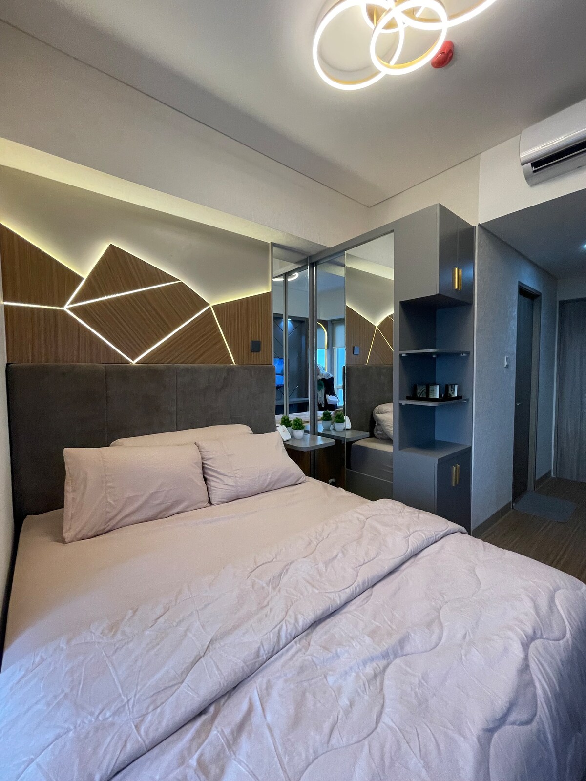 A sleeping area features a spacious bed with soft bedding and contemporary wall accents that incorporate geometric lighting. A mirror reflects the room, enhancing the sense of space, while a minimalist shelving unit is positioned nearby. Natural light is allowed in through a window.