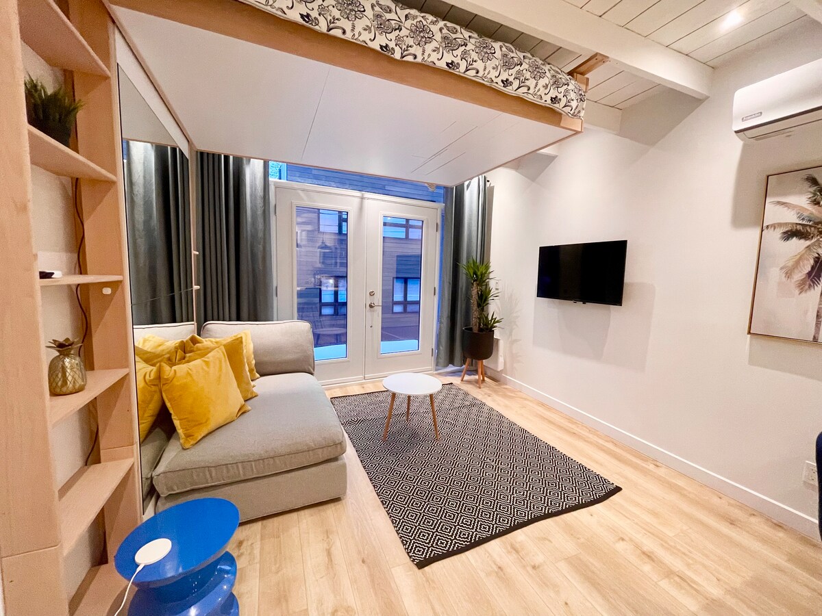 A light-filled living area features a comfortable gray sofa adorned with yellow cushions. A coffee table is placed on a patterned area rug, and a flat-screen TV is mounted on the wall. Large windows with dark curtains open to a balcony space.