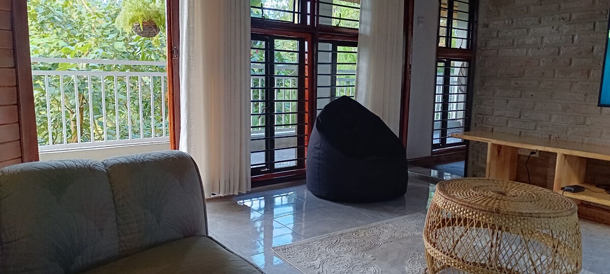 A spacious living area is illuminated by natural light from large windows. A plush black bean bag is positioned on polished tile flooring. A circular woven coffee table sits nearby, accompanied by a soft area rug. Light-colored curtains frame the windows, enhancing the inviting atmosphere.