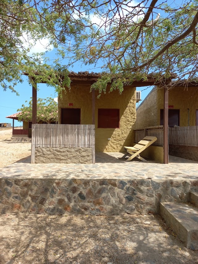 A rustic two-unit structure is framed by low stone walls and shaded by trees. Each unit features wooden shutters and a simple porch area, with a wooden lounge chair positioned for relaxation. The landscape showcases dry terrain under a clear blue sky.