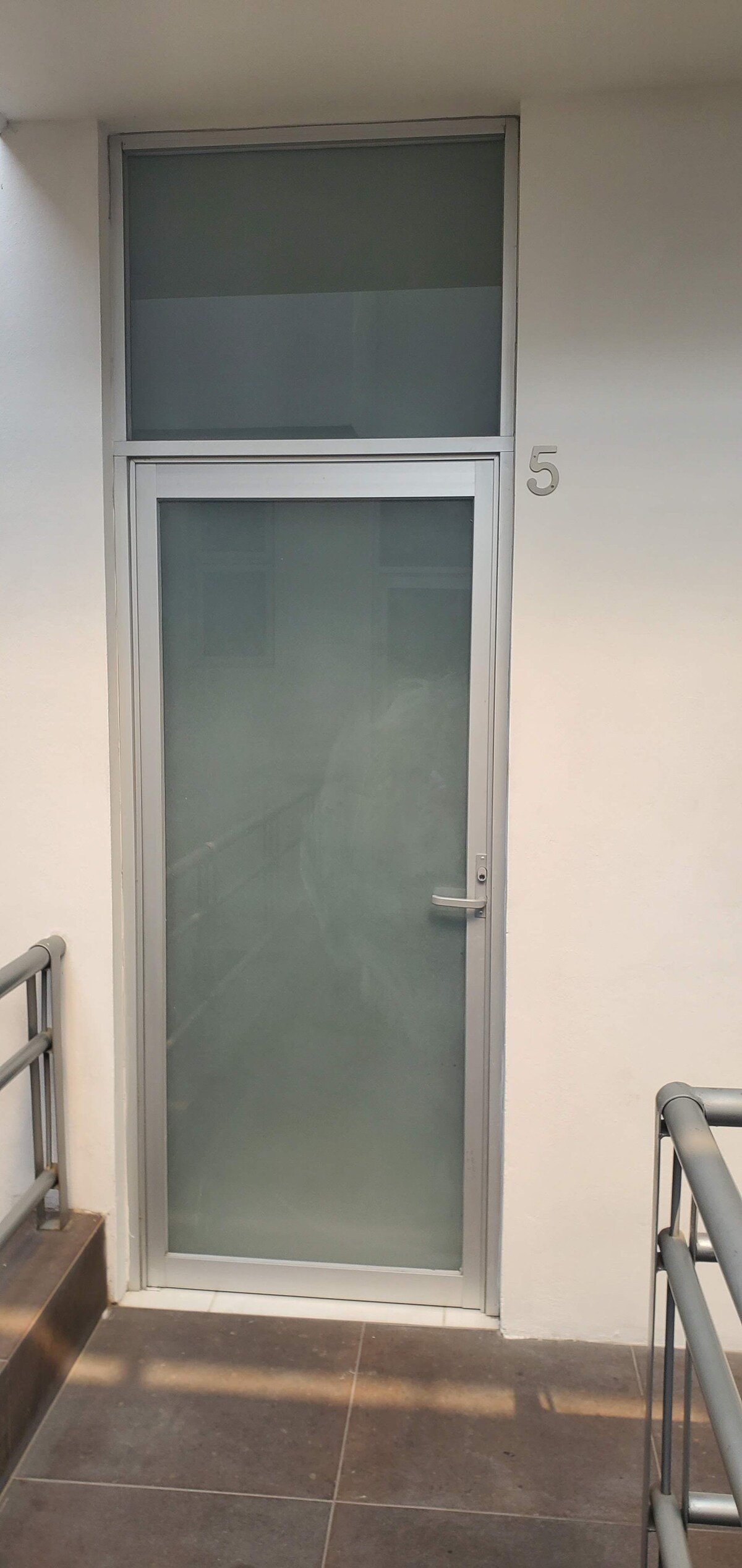 A sleek glass door marked with the number 5 is positioned at the entrance. The door features a frosted design that offers privacy, and is framed by a simple metal border. Light beige walls complement the minimalistic aesthetic.
