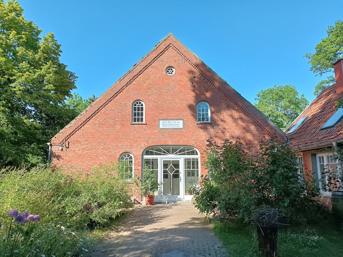 A charming brick structure features a distinctive peaked roof and large front windows that invite natural light indoors. The entrance is framed by greenery, with flower beds and pathways leading to the door, creating a welcoming ambiance.