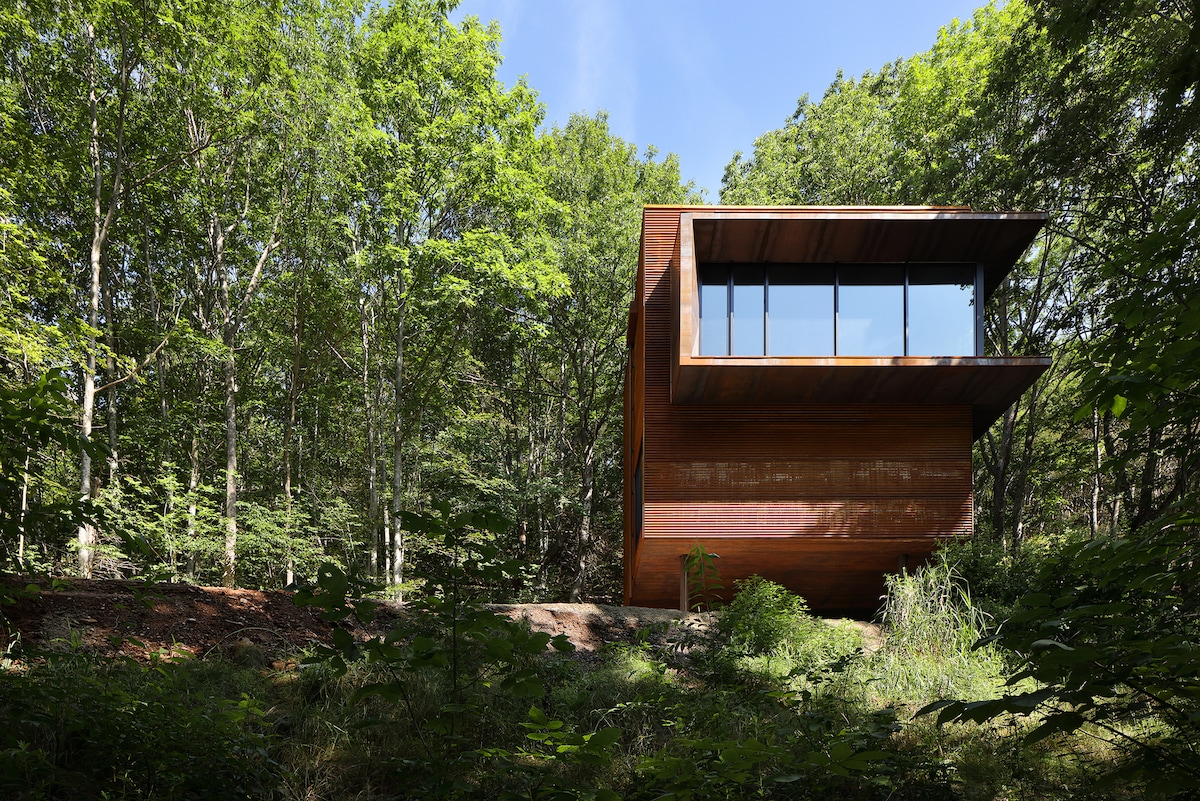 A contemporary structure with a wooden exterior is nestled among lush greenery. Large glass windows offer unobstructed views of the surrounding forest, while a cantilevered roof creates a distinct architectural feature. The natural landscape frames the home, enhancing the serene and secluded setting.