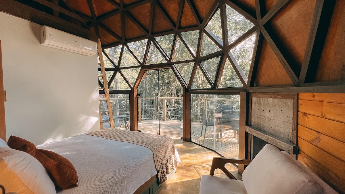 The interior of the geodesic dome features a spacious bed and a comfortable chair. Large triangular windows provide ample natural light and a view of the surrounding nature. A wooden ladder leads to an elevated area, enhancing the unique architectural design.