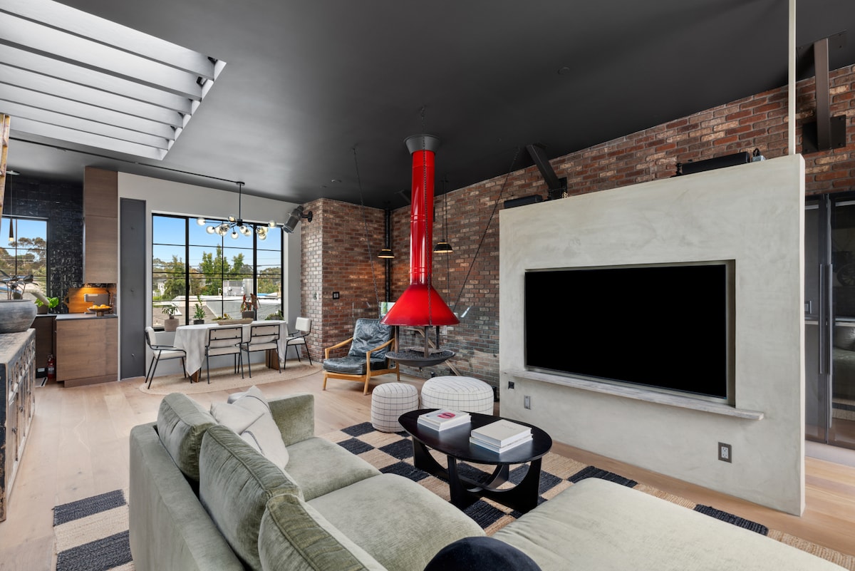 An open-concept living area features a modern layout, combining natural textures with sleek design. Exposed brick walls provide warmth against the cool tones of the furnishings. A striking red fireplace serves as a focal point, while large windows allow ample natural light.