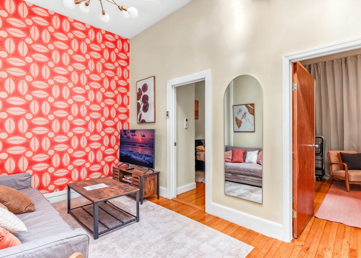 A living area is showcased with a large couch adorned with decorative pillows, accompanied by a coffee table. An abstract wall art piece complements the vibrant orange accent wall. A TV is positioned in front of the couch, while a mirror adds depth to the space.