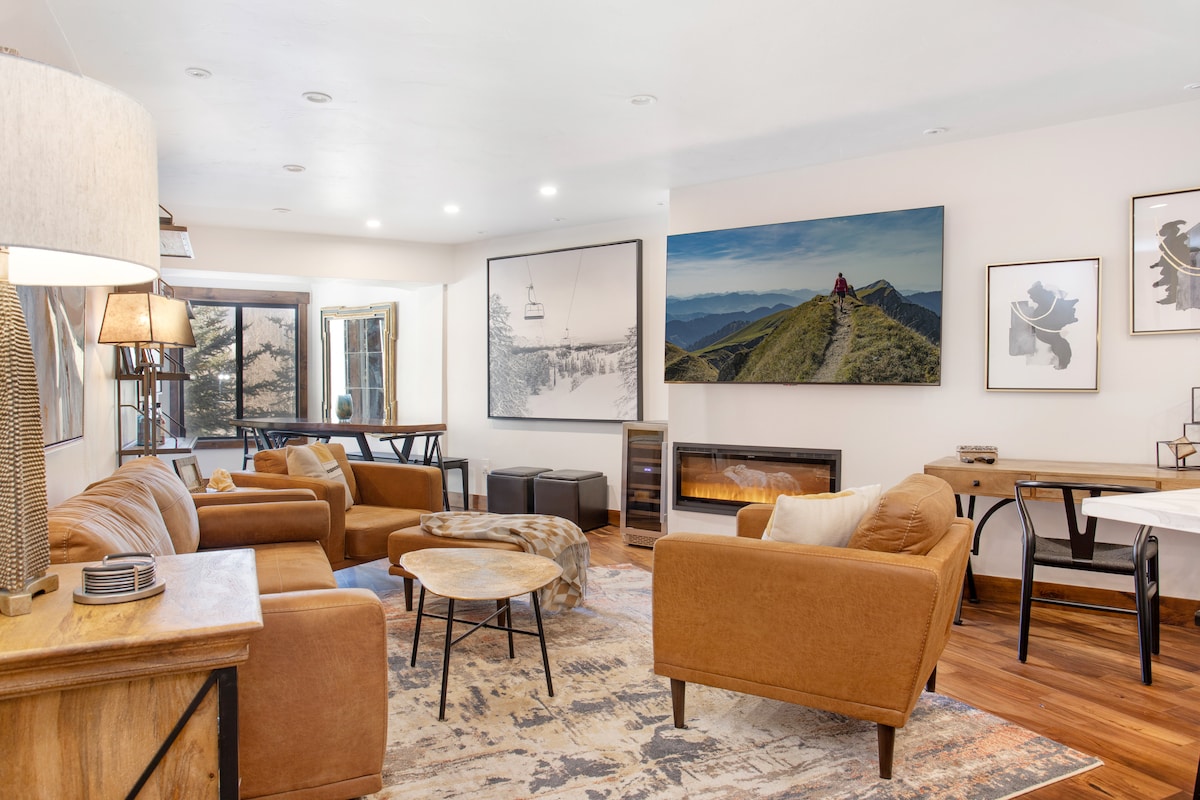The living area features ample seating arranged around a round coffee table, complemented by a gas fireplace. A large artwork depicting a mountainous landscape adorns one wall, while floor-to-ceiling windows allow for natural light and views of the surrounding landscape.