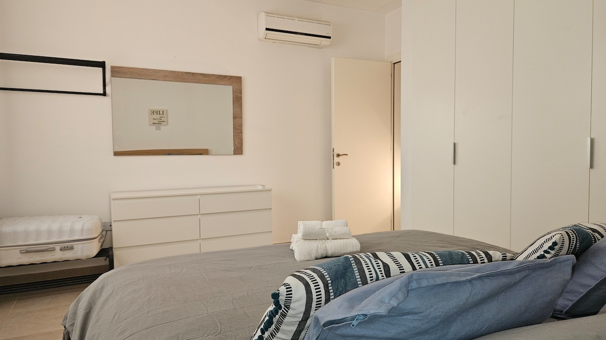 A spacious bedroom is presented with a comfortable bed adorned with decorative pillows. A large mirror reflects natural light, while a dresser and a hanging rack provide ample storage. A compact bed for children is positioned in the corner, contributing to the room's family-friendly layout.
