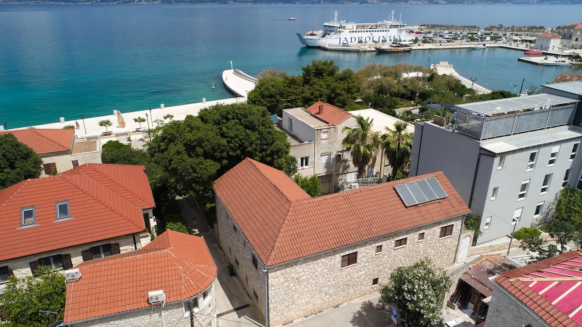 A scenic view of the seaside area in Supetar showcases the water's calm surface alongside the harbor. Traditional stone buildings with red roofs complement the lush greenery. A ferry can be seen docked, while the shoreline offers easy access to the waterfront.