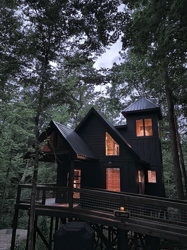 A modern treehouse is surrounded by lush green trees, featuring a dark exterior with multiple peaked roofs. Soft illumination radiates from the windows, highlighting the structure's unique architectural design. The elevated deck is visible, providing a connection to the forested environment.