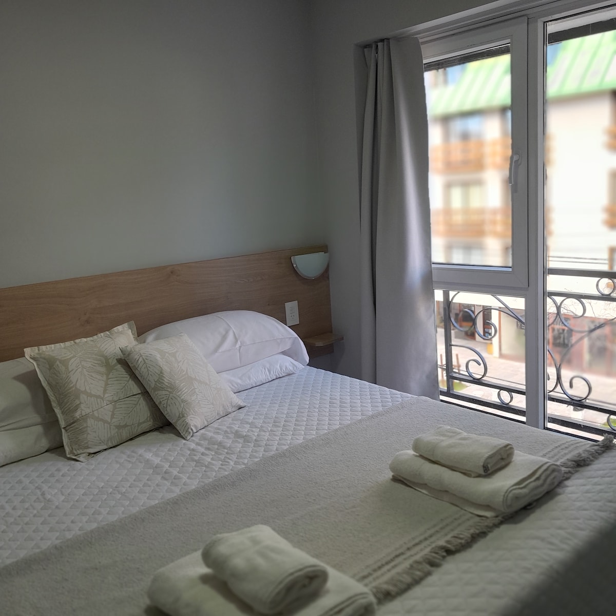 A cozy sleeping area is furnished with a spacious bed topped with neutral-toned bedding and decorative pillows. Soft towels are neatly arranged at the foot of the bed. A large window allows natural light in, showcasing a view of the outside with a decorative balcony railing.