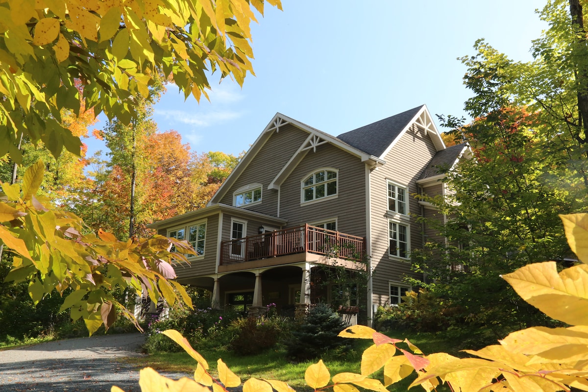 The chalet is surrounded by vibrant autumn foliage, featuring a well-maintained exterior with large windows and decorative gables. A spacious deck with railings is visible, offering an inviting entrance. The landscape is enhanced by mature trees and a gravel pathway leading to the front.