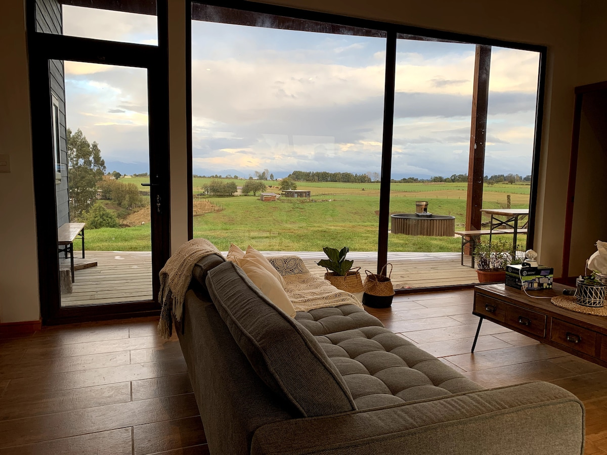 A spacious living area features a comfortable sofa facing large glass doors that open to a scenic view of green fields and distant mountains. Natural light fills the room, highlighting the wooden floor and tasteful decor elements, including a coffee table and decorative plants.
