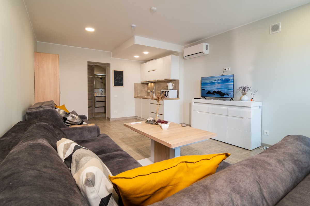 A spacious living area is designed with a comfortable sectional sofa featuring yellow and gray accent pillows. A wooden coffee table sits in front, with a view of a flat-screen TV against a light-colored wall. The open kitchen area includes modern appliances, creating a welcoming atmosphere.