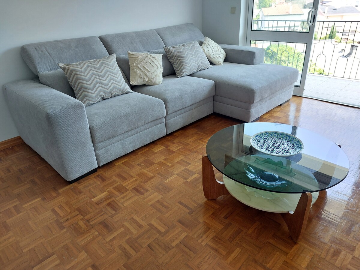A modern sectional sofa in a soft gray fabric is accompanied by textured cushions. A contemporary glass coffee table with a unique design sits on a parquet floor, showcasing a decorative bowl at its center. Natural light brightens the space through an adjacent sliding door.