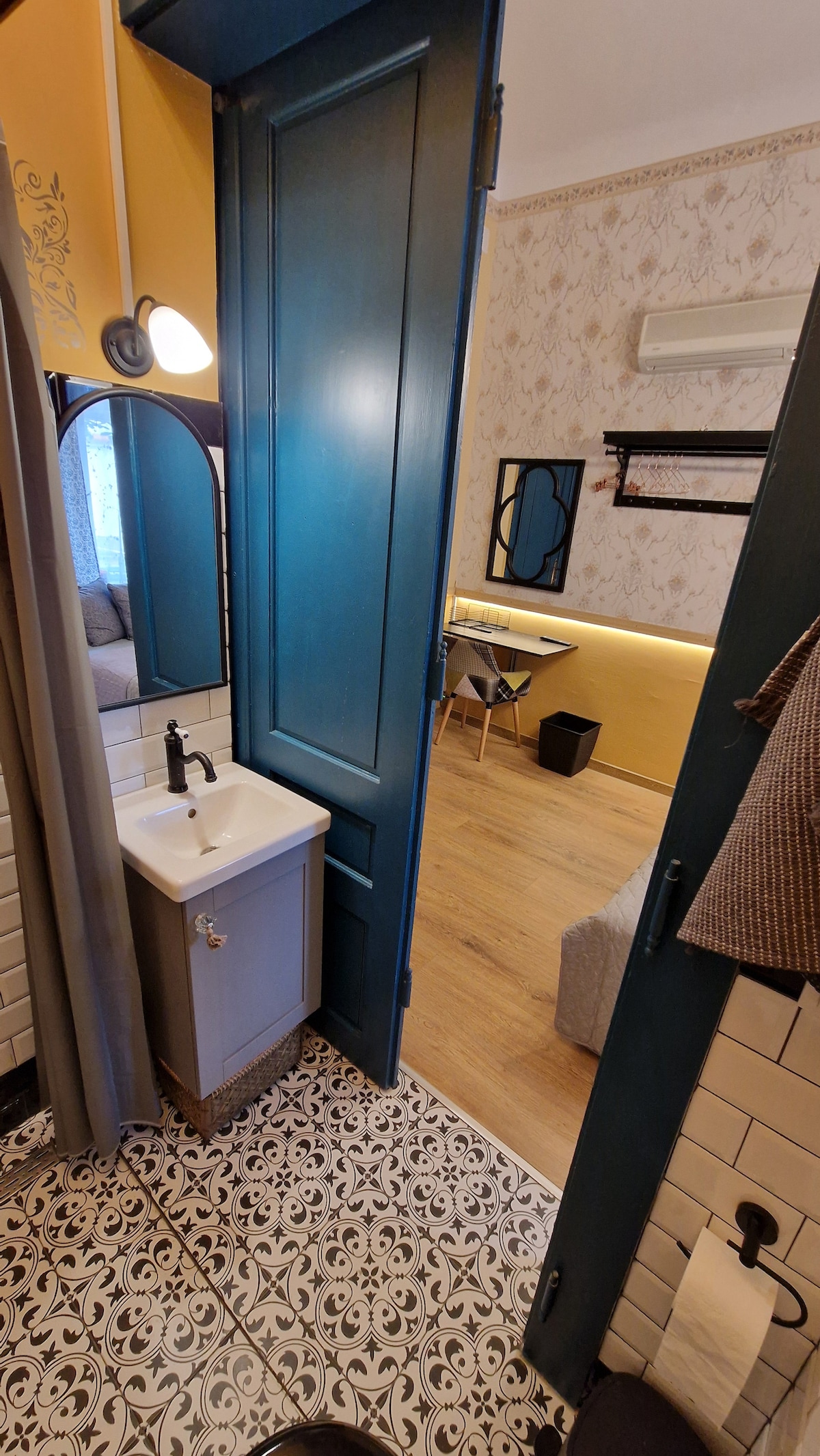 A glimpse into a well-designed bathroom with patterned tiles and a small sink. A blue door leads into a softly lit room featuring a cozy seating area with a gray couch and a desk. Light wood flooring enhances the overall inviting atmosphere.