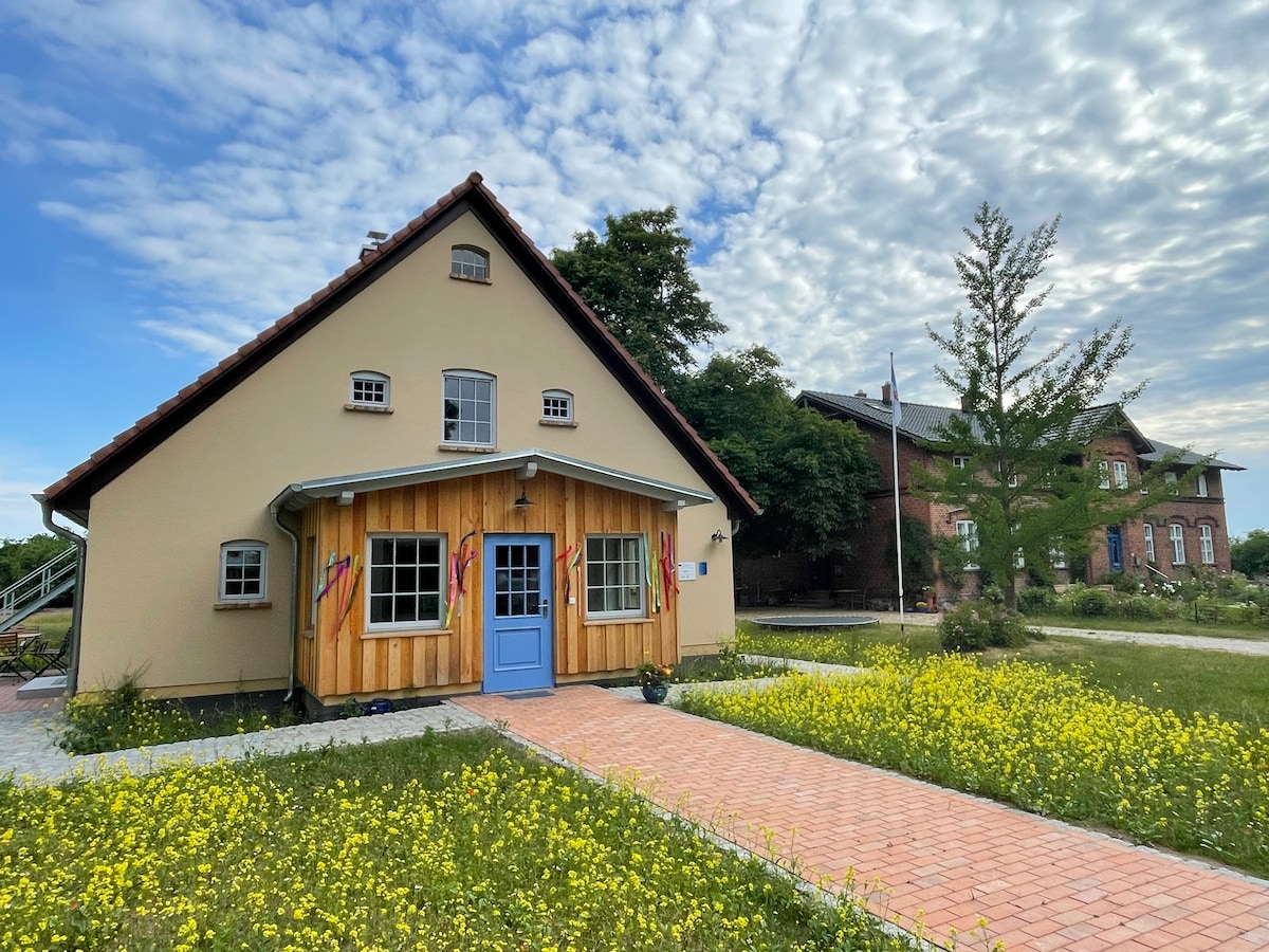 A welcoming building features a mix of wood and stucco exteriors with a distinctive blue door at the front. Colorful flags adorn the entrance, bordered by a brick pathway leading through a meadow of yellow flowers. The sky shows scattered clouds, complementing the serene landscape.