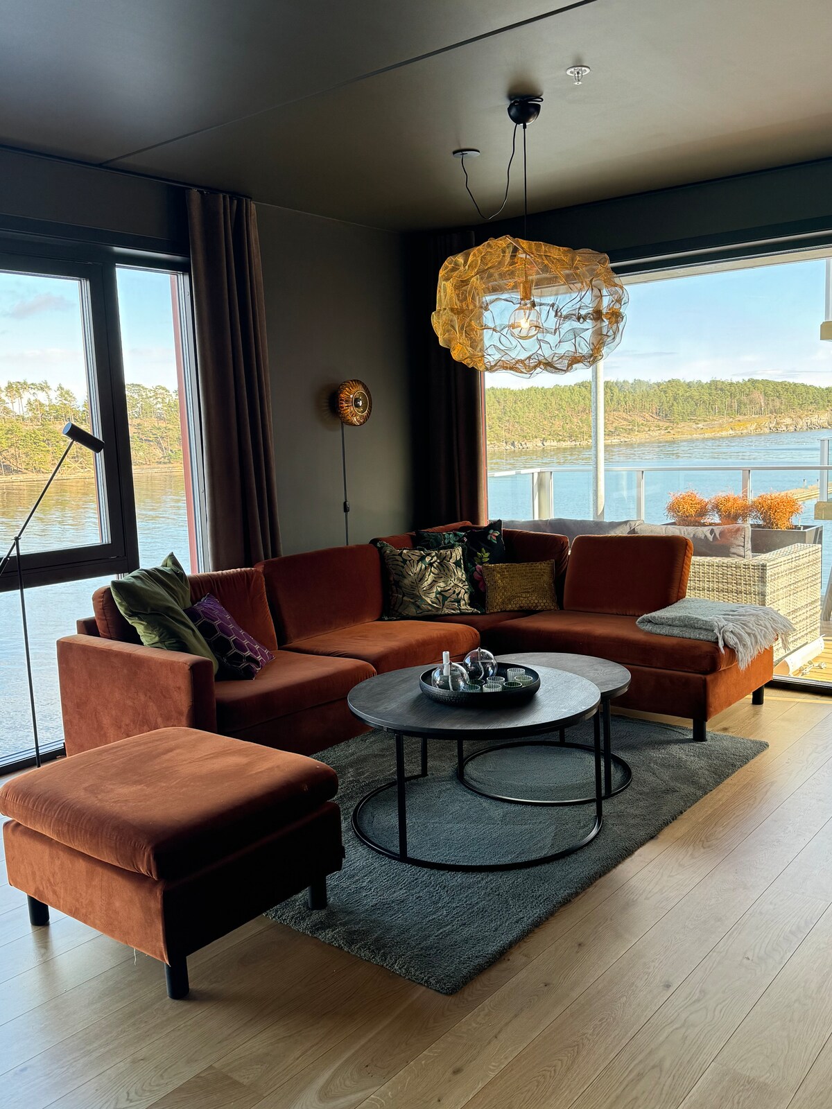 A comfortable living area is highlighted by a plush sofa and an armchair, complemented by a round coffee table. Large windows offer views of the serene waters, while natural light enhances the warm tones of the furnishings. A decorative light fixture adds a touch of style.