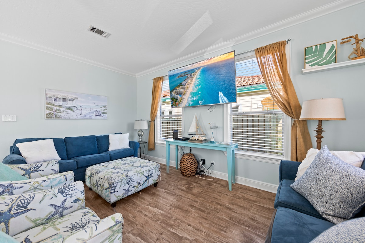 A cozy living area is showcased with blue sofas and a patterned armchair, complemented by decorative pillows. A large screen television is mounted on the wall, while light curtains frame the windows, allowing natural light to brighten the space. A small table with beach-themed decor enhances the coastal ambiance.